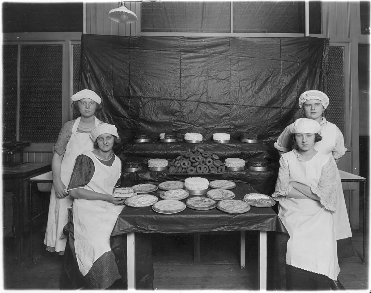 Black-and-white photo of students in aprons and caps with baked goods at a vocational school exhibit